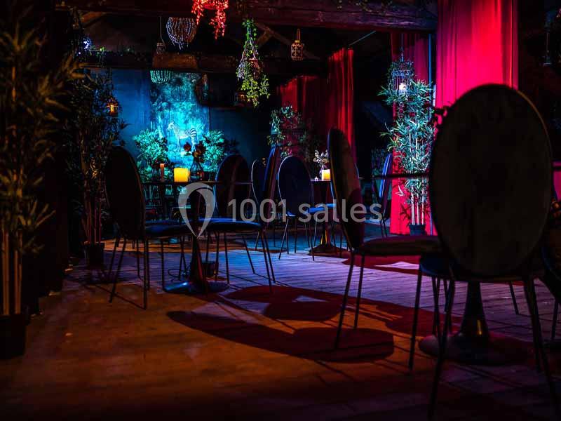 Salle sombre avec tables et chaises, éclairée par des bougies et des lumières colorées, entourée de plantes et rideaux…