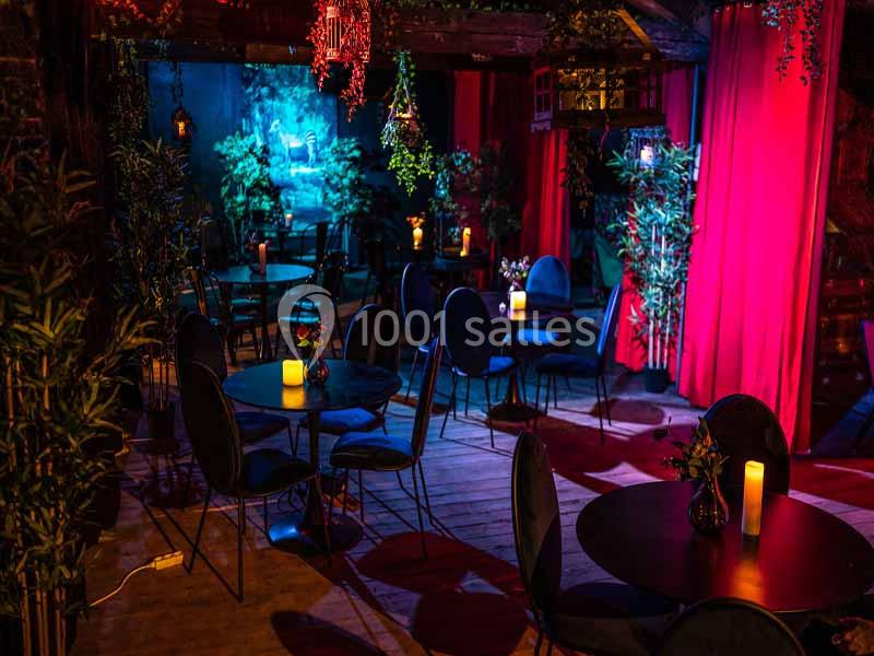 Salle sombre avec tables rondes, chaises noires, bougies allumées et rideaux rouges, décorée de plantes et lumières tamisées.