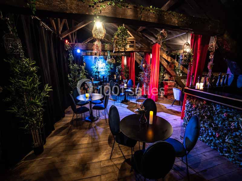 Salle décorée avec des tables rondes, chaises noires, lumières tamisées, rideaux rouges et végétation suspendue.