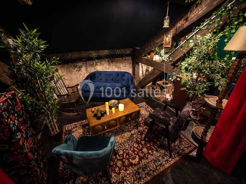 Salon chaleureux sous combles avec canapé bleu, fauteuils, tapis persan, plantes et éclairage tamisé.