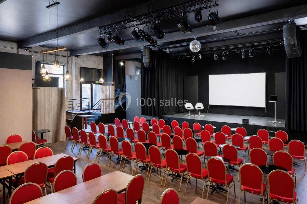 Salle de conférence avec des rangées de chaises rouges, une scène équipée d'un écran blanc et un éclairage suspendu.