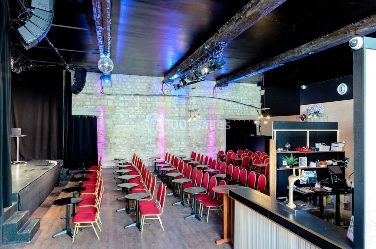 Salle de spectacle avec rangées de sièges rouges, petites tables noires, scène à gauche et mur en pierre apparente.