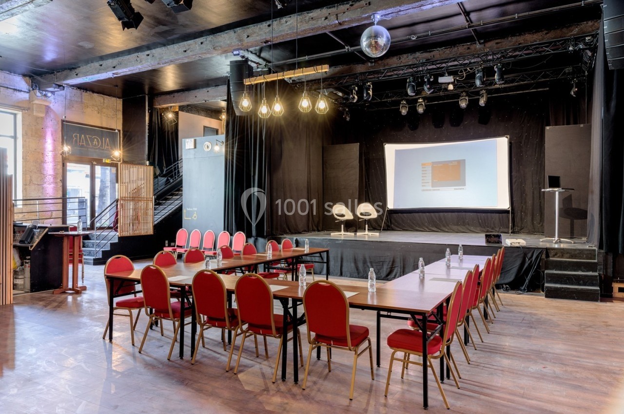 Salle de conférence avec scène, deux fauteuils, écran de projection et tables disposées en U avec chaises rouges.