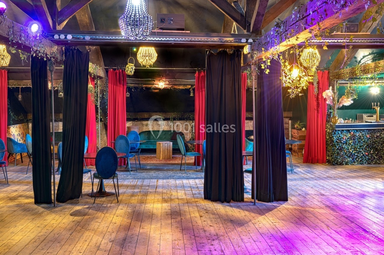 Salle avec parquet en bois, rideaux rouges, chaises bleues, éclairage chaleureux et bar décoré de végétation.