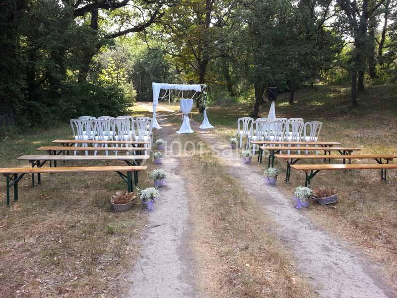 Disposition de bancs et chaises en extérieur, menant à une arche décorée pour une cérémonie dans un cadre boisé.