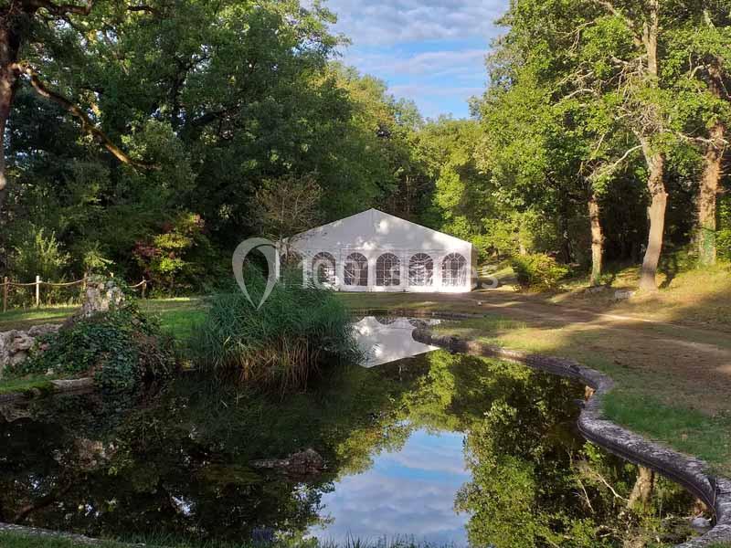 Tente blanche installée dans un espace naturel verdoyant, près d'un étang reflétant les arbres environnants.