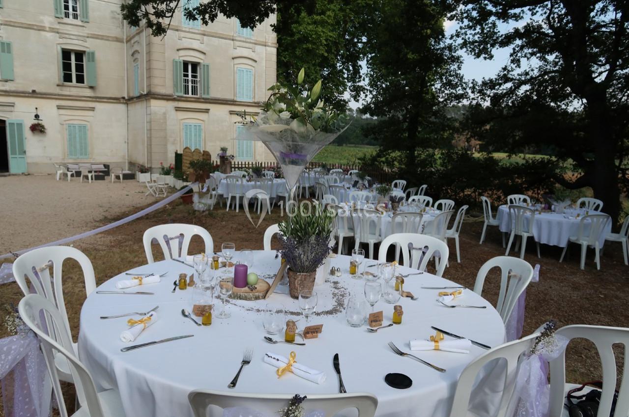 Table de réception extérieure décorée avec des fleurs et des rubans, devant un bâtiment ancien et un jardin arboré.