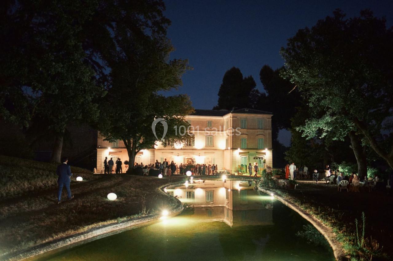 Manoir éclairé de nuit, entouré d'arbres, avec un étang réfléchissant les lumières et des invités rassemblés.