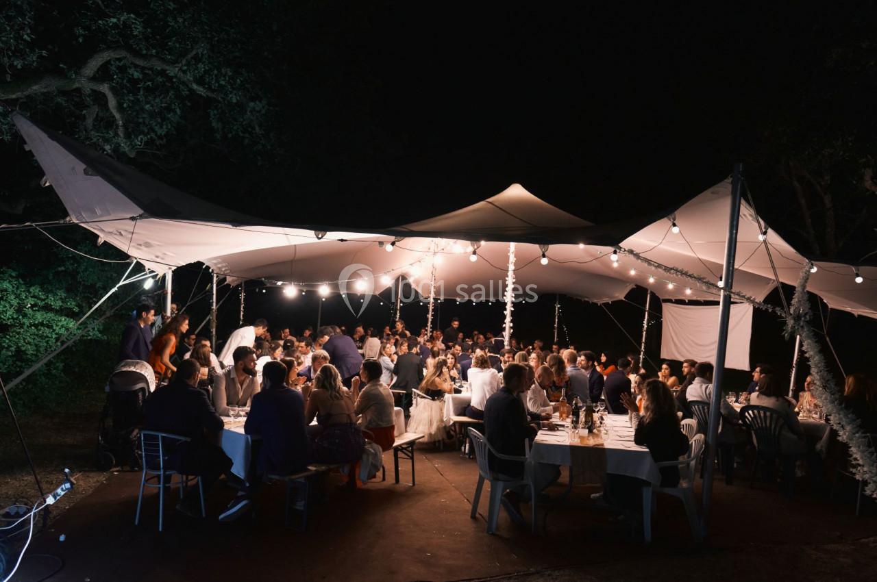 Des convives dînent sous une tente éclairée par des guirlandes lumineuses, en pleine nuit, dans un cadre extérieur.