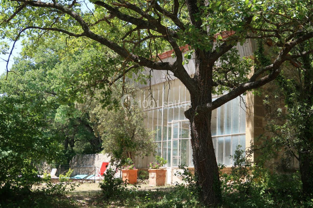 Serre en verre adossée à un bâtiment en pierre, entourée de végétation et d'arbres sous un ciel dégagé.