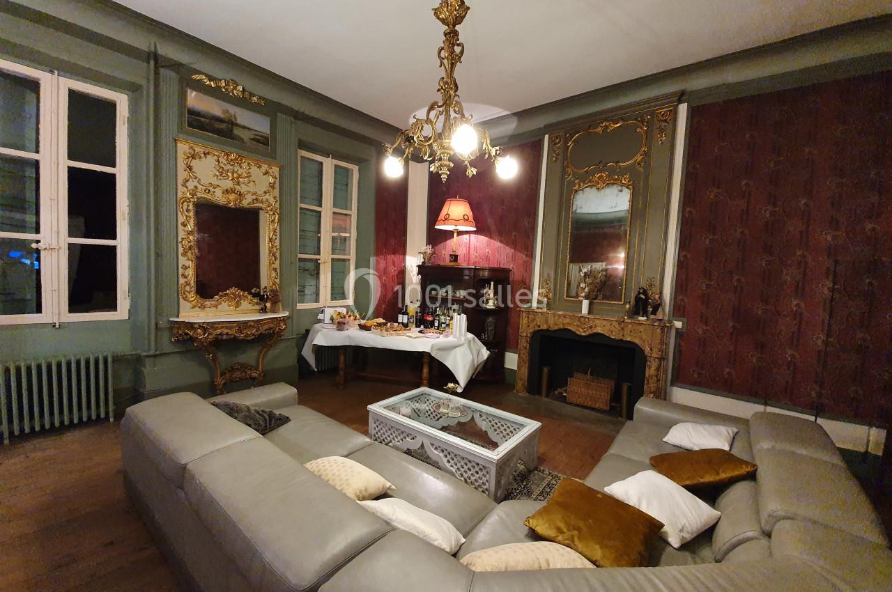 Salon ancien avec moulures dorées, canapé d'angle gris, cheminée, lustre et table dressée pour un buffet.