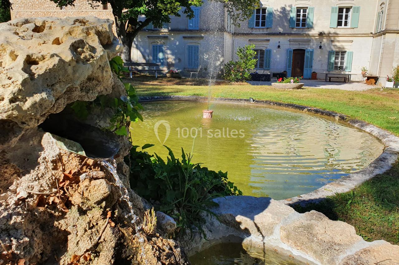 Fontaine en pierre avec un jet d'eau au centre d'un bassin rond, devant un bâtiment ancien entouré de verdure.
