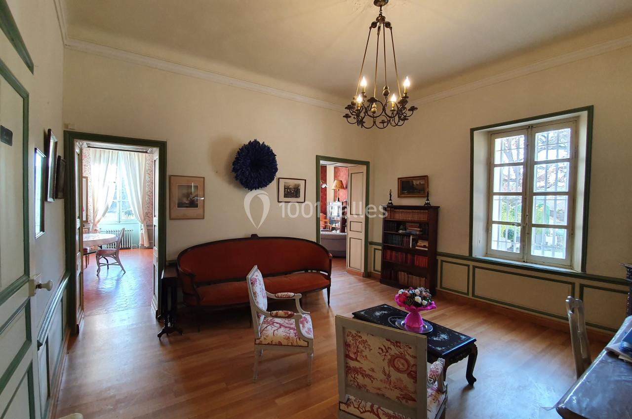 Salon lumineux avec parquet, canapé, fauteuils, bibliothèque, lustre et vue sur une pièce adjacente éclairée.