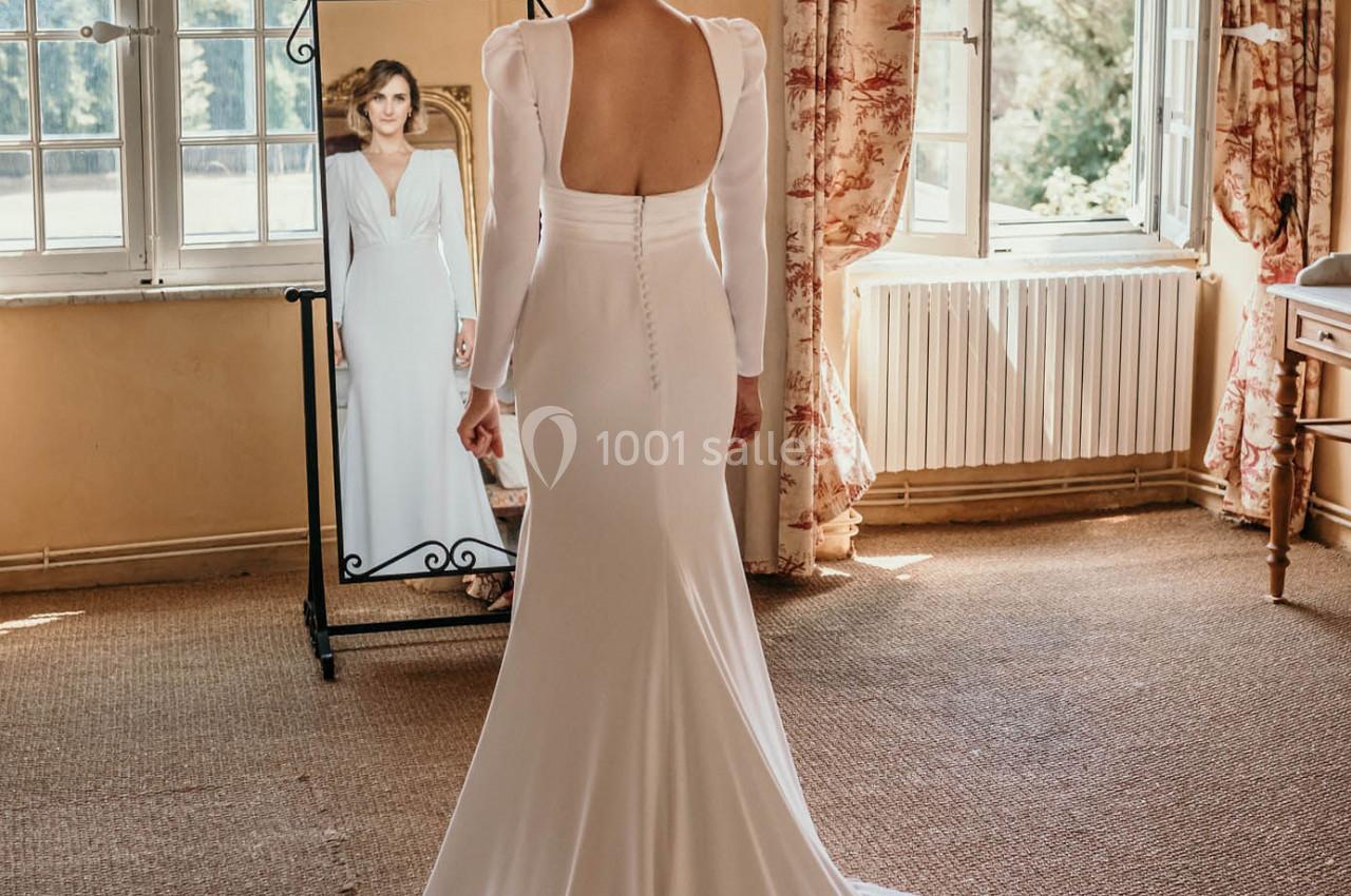 Une femme en robe de mariée blanche se regarde dans un miroir dans une pièce lumineuse avec de grandes fenêtres.