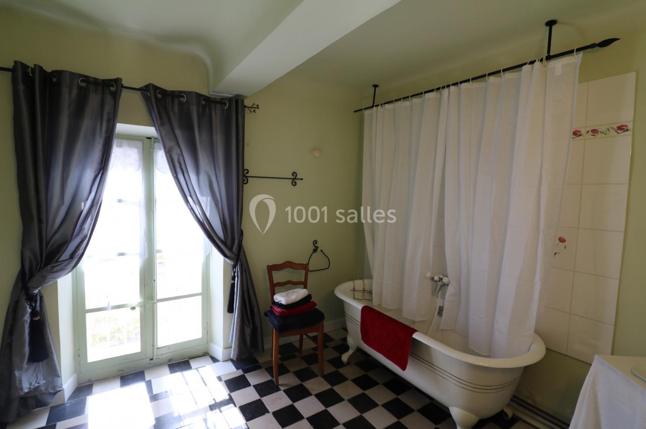 Salle de bain avec baignoire sur pieds, rideau de douche blanc, carrelage noir et blanc, et fenêtre avec rideaux gris.