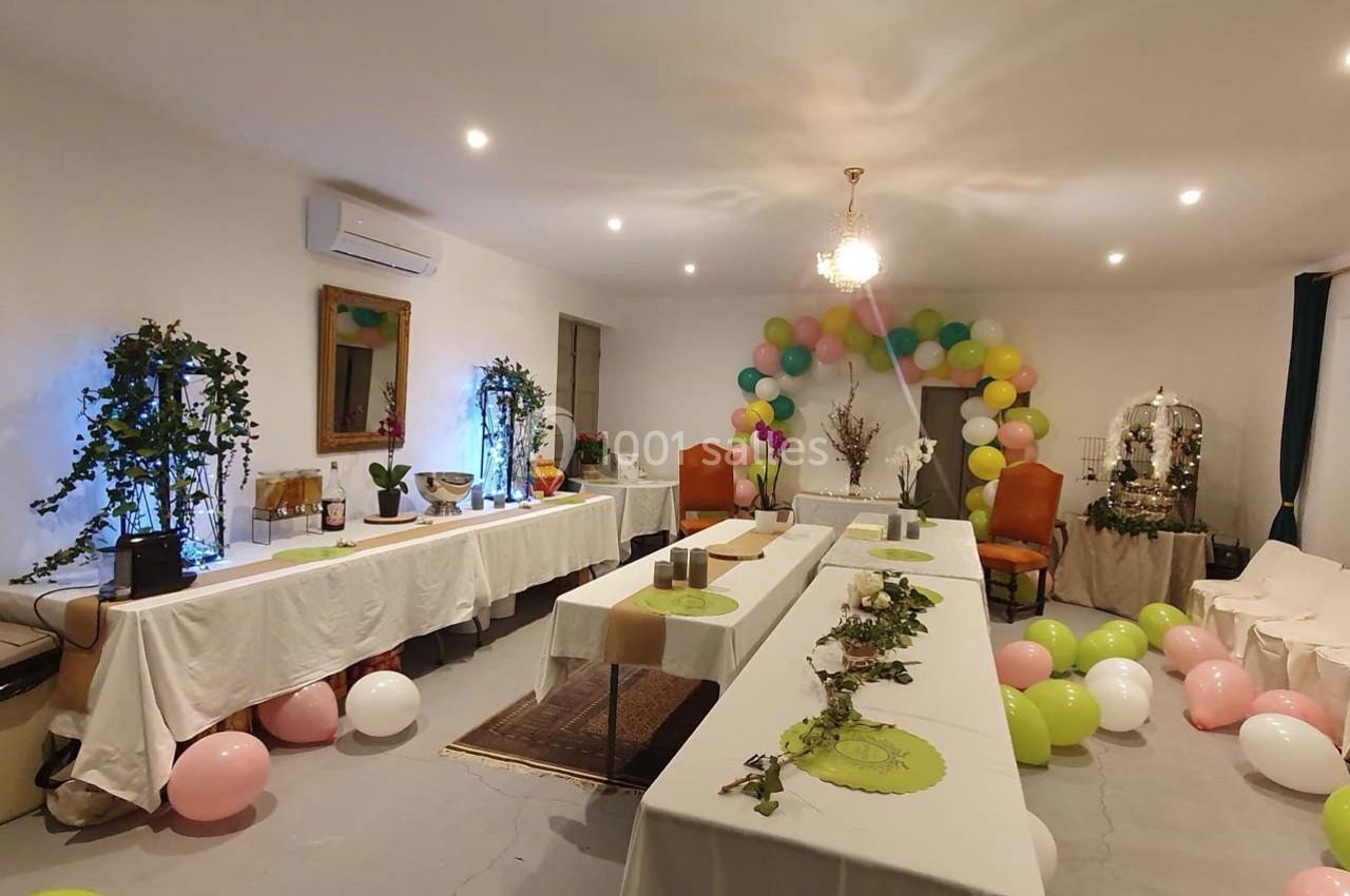Salle décorée pour une réception avec ballons colorés, guirlandes végétales et tables couvertes de nappes blanches.