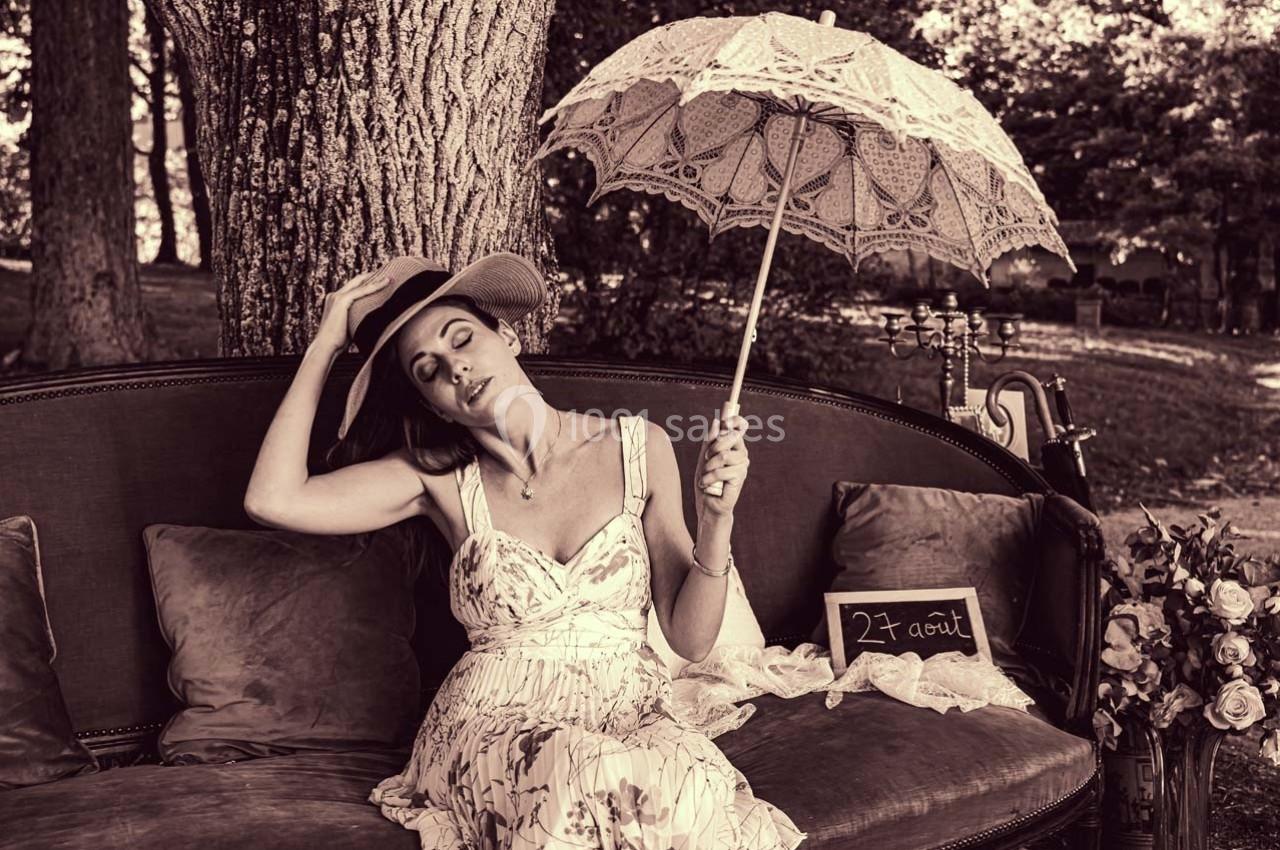 Femme assise sur un canapé vintage, tenant une ombrelle décorative, dans un parc ombragé.