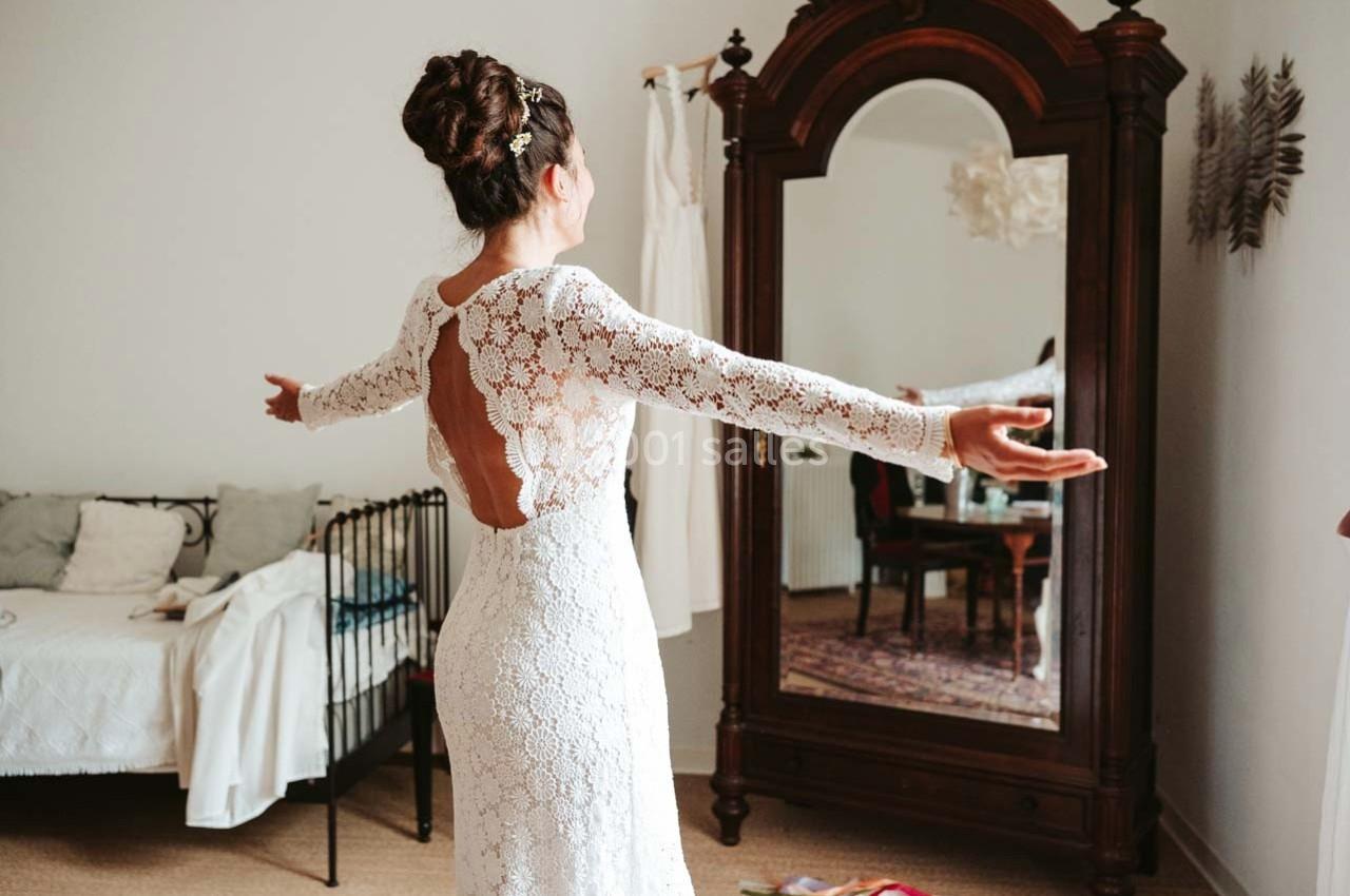 Femme en robe de mariée en dentelle blanche, bras ouverts, devant un miroir ancien dans une chambre lumineuse.