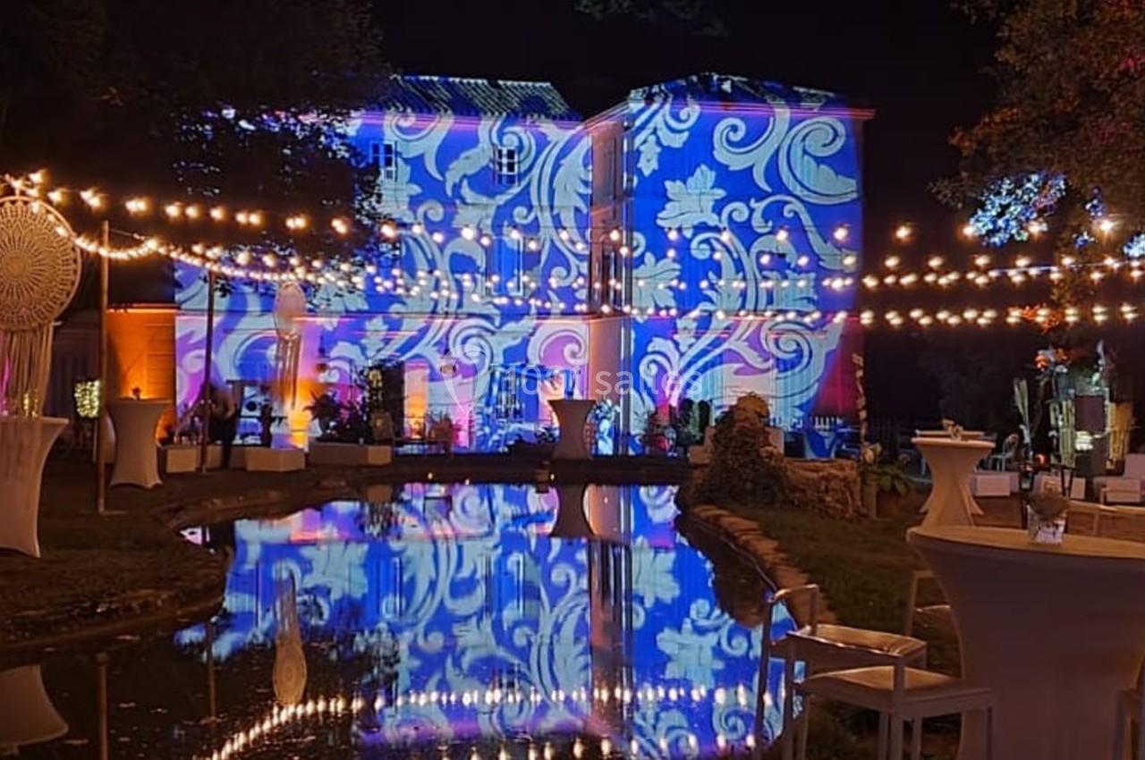 Façade d'un bâtiment illuminée par des motifs colorés, avec guirlandes lumineuses et leur reflet dans un plan d'eau.