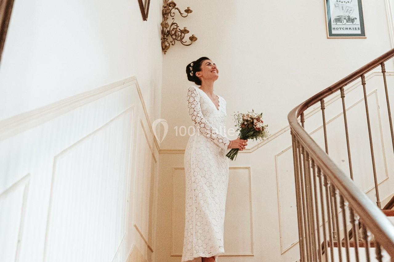 Une femme en robe blanche tient un bouquet de fleurs en haut d'un escalier dans un intérieur lumineux.