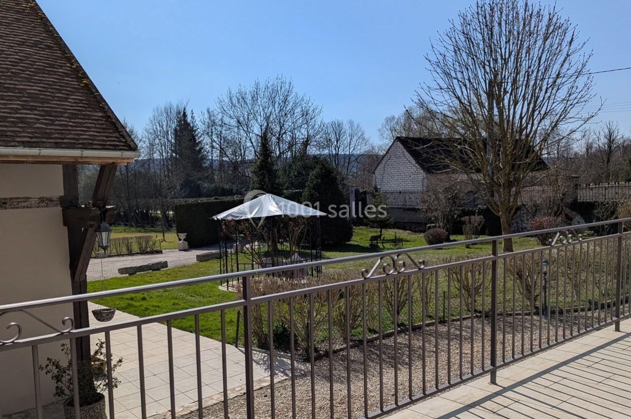 Vue d'un jardin avec pelouse, arbres, pergola métallique et bâtiments en arrière-plan, prise depuis une terrasse.