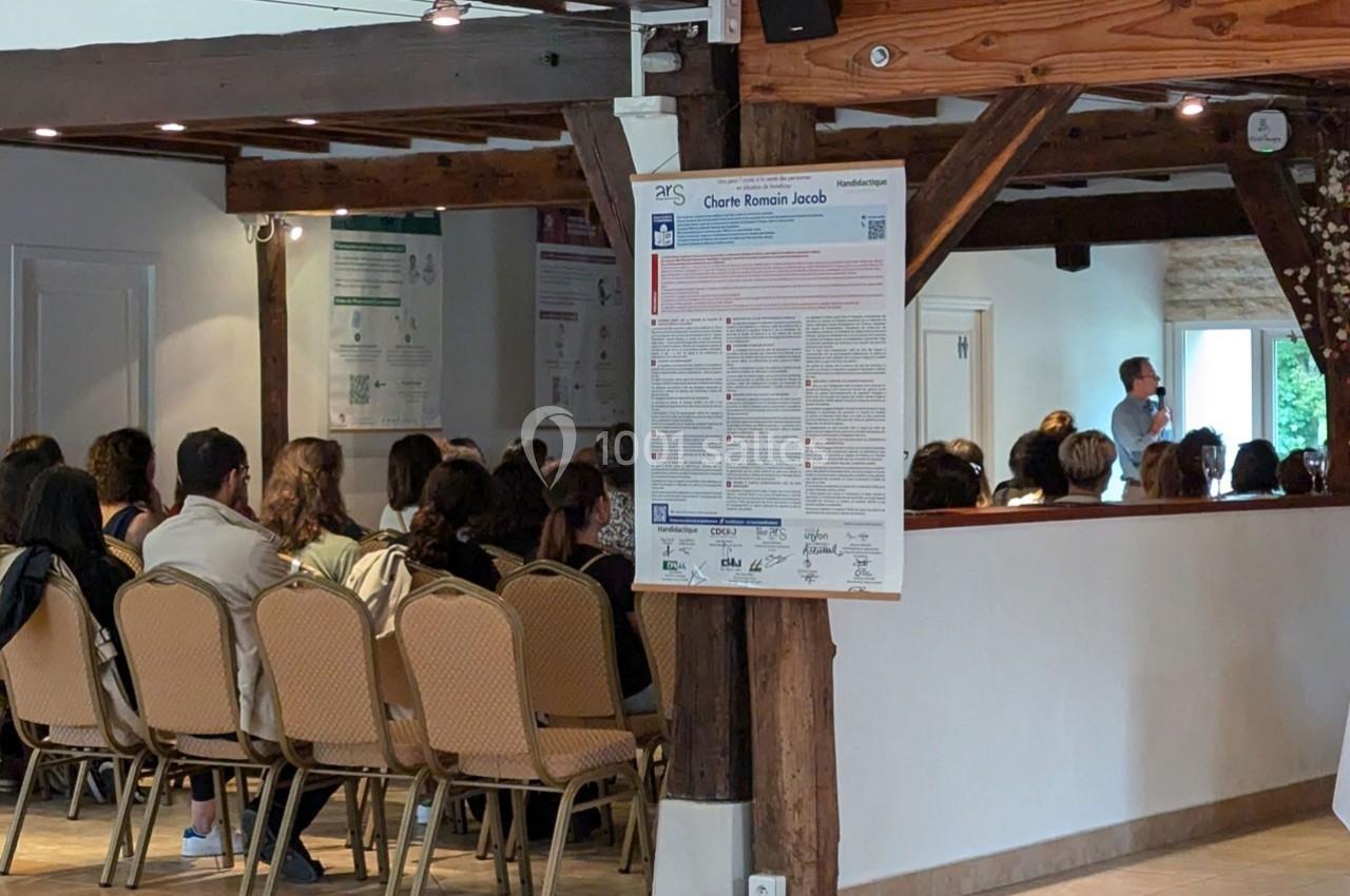 Un groupe de personnes assises dans une salle, écoutant un intervenant debout près d'un mur en bois.