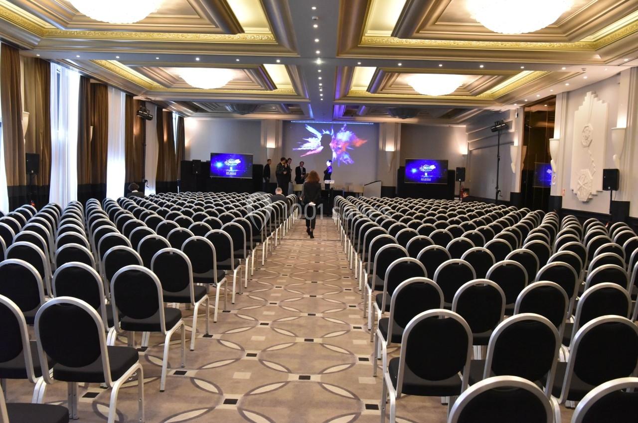 Salle de conférence vide avec des rangées de chaises noires, un écran et une scène éclairée en arrière-plan.