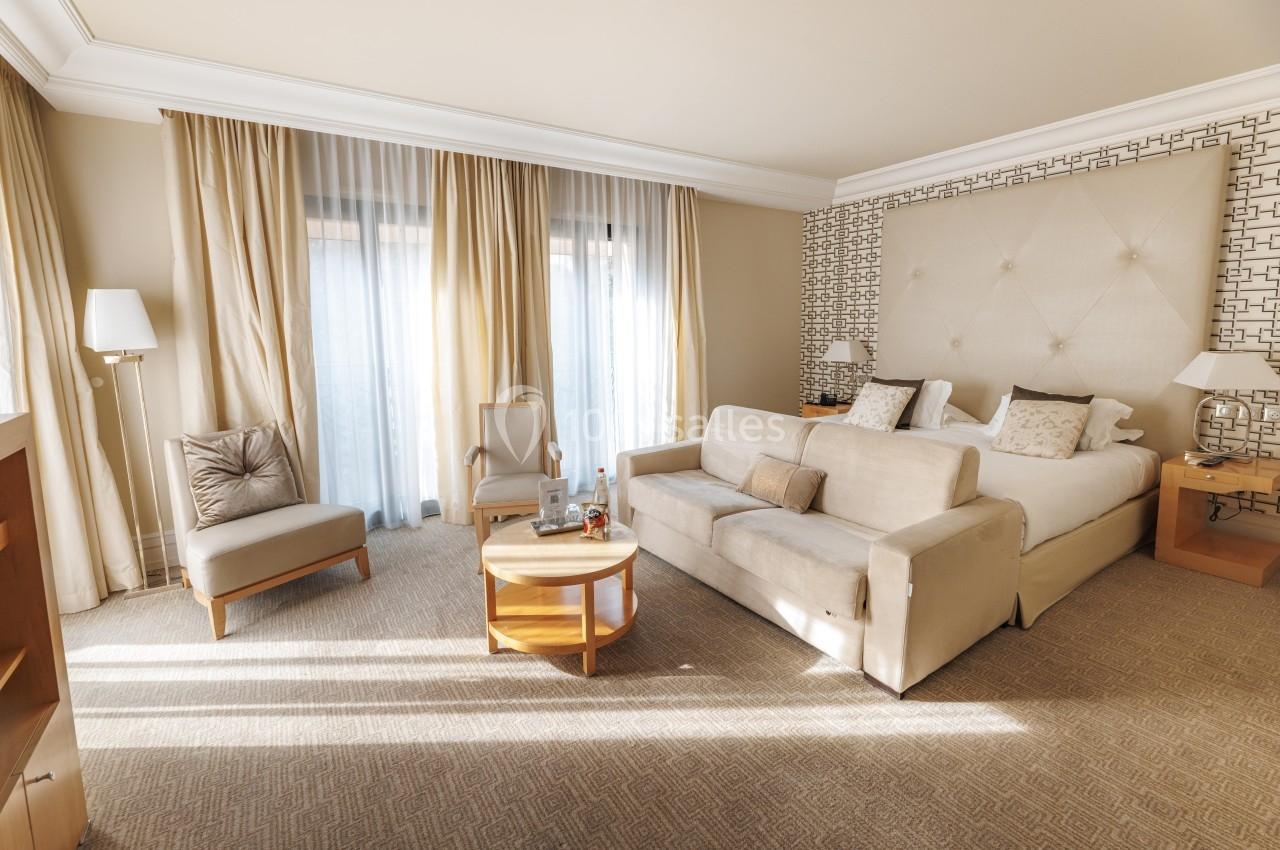 Chambre lumineuse avec lit double, canapé, fauteuils, table basse et grandes fenêtres avec rideaux beiges.