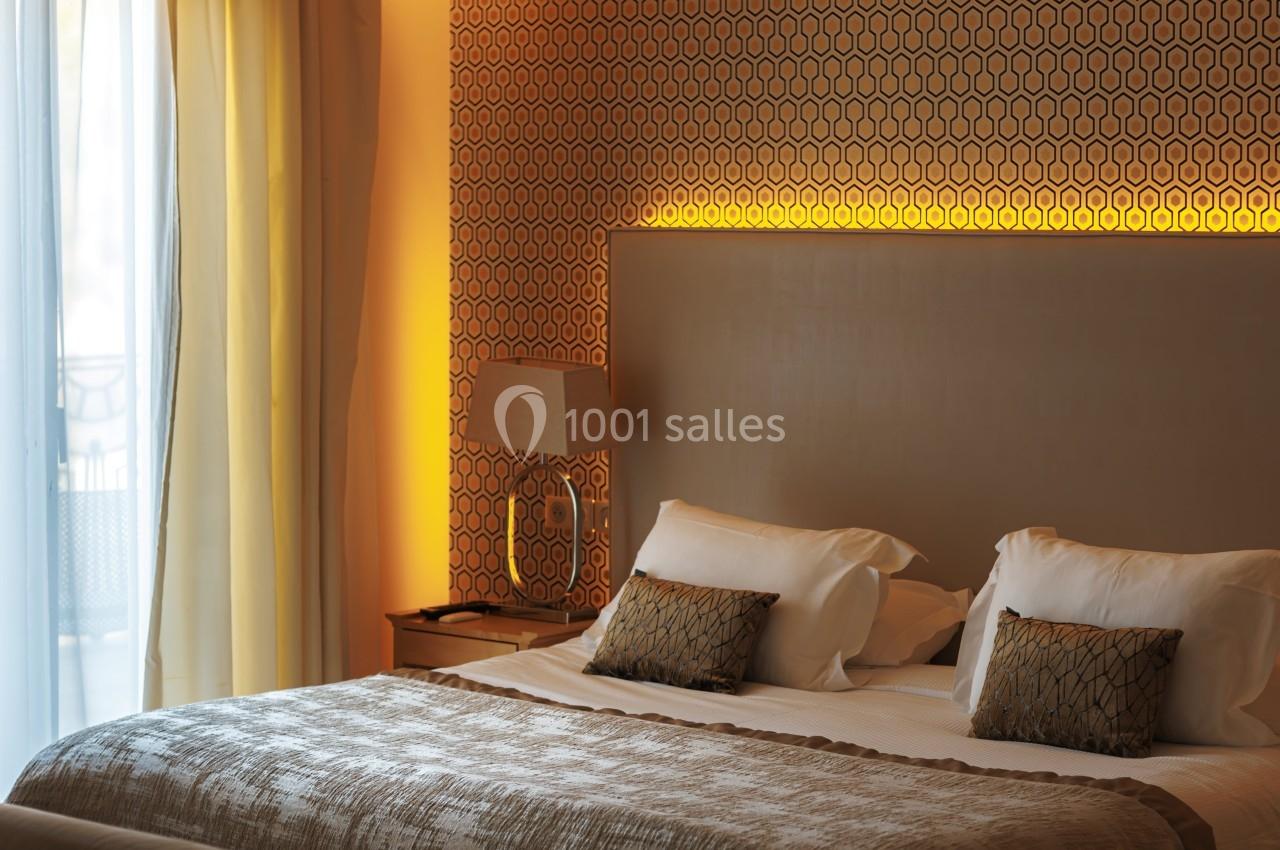 Lit double avec coussins décoratifs, lampe de chevet et éclairage tamisé, près d'une fenêtre avec rideaux clairs.