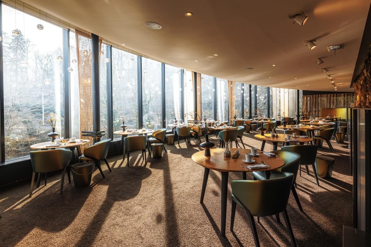 Salle de restaurant lumineuse avec de grandes baies vitrées, tables en bois et chaises modernes.