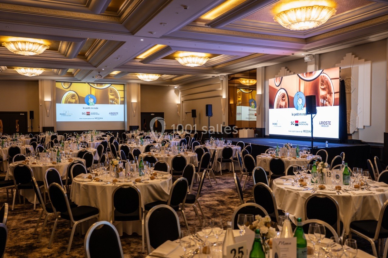 Salle de réception élégante avec tables dressées, écrans de projection et éclairage chaleureux au plafond.