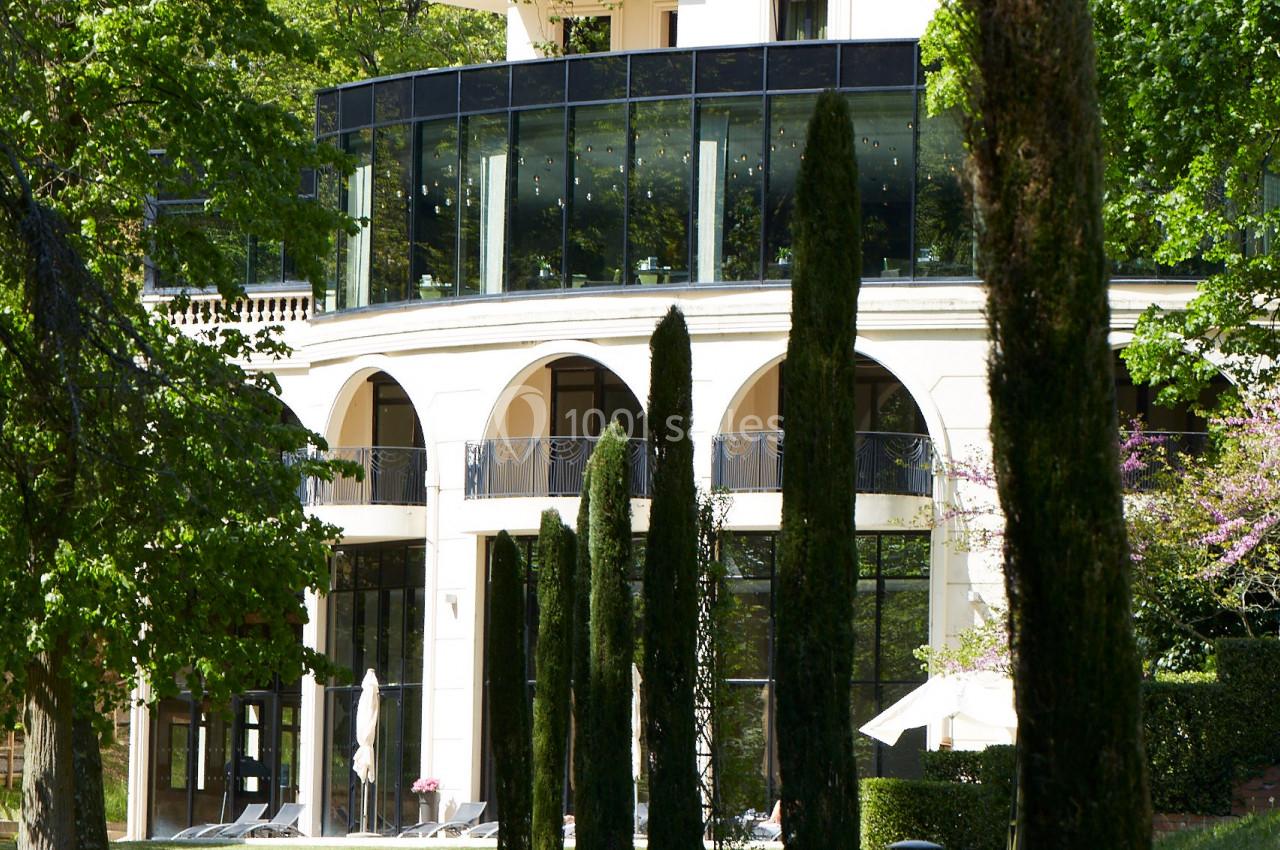 Façade d'un bâtiment moderne entouré d'arbres et de cyprès, avec une pelouse au premier plan.
