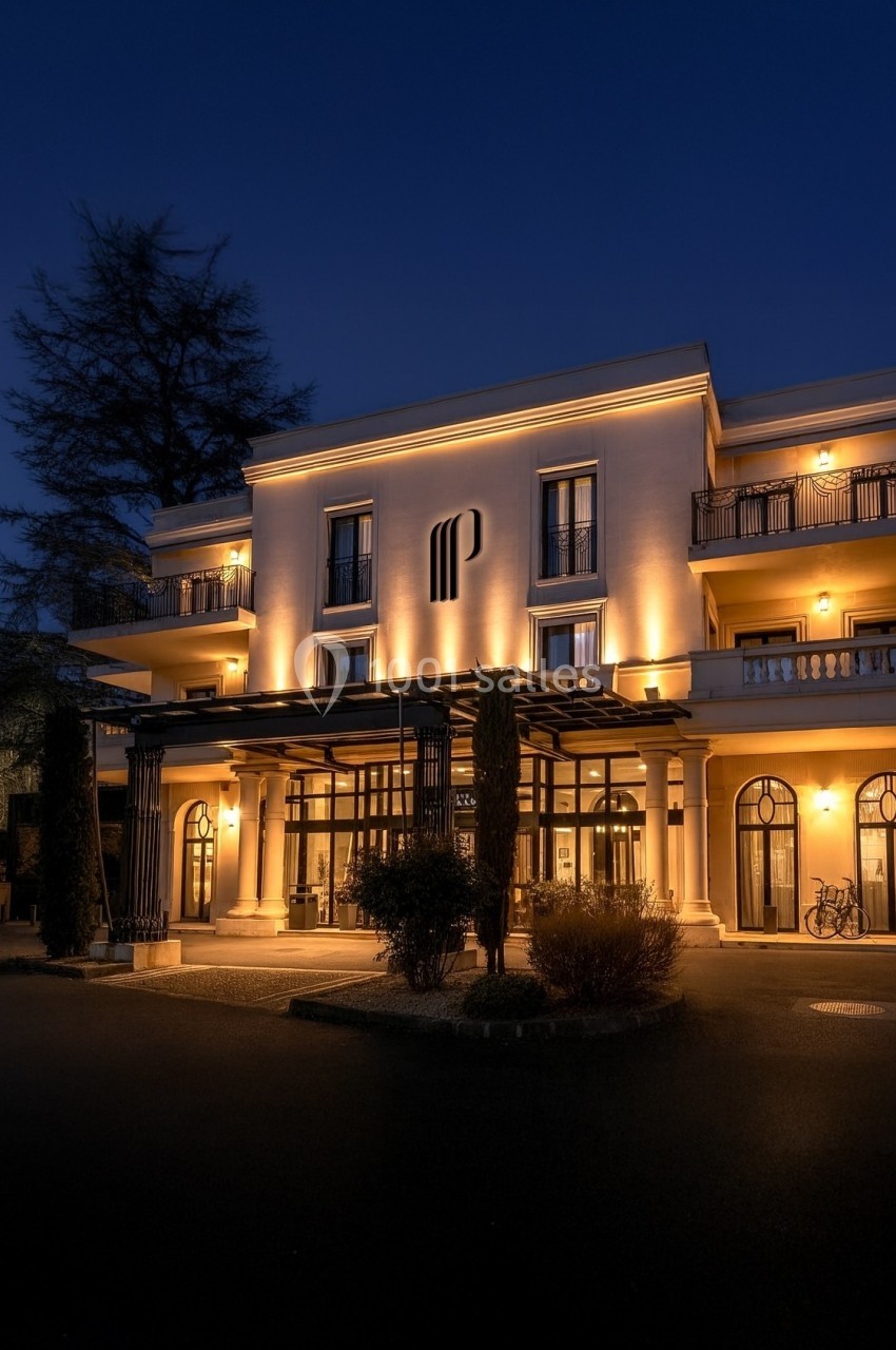 Façade éclairée d'un bâtiment élégant à trois étages, entouré d'arbres, photographié à la tombée de la nuit.