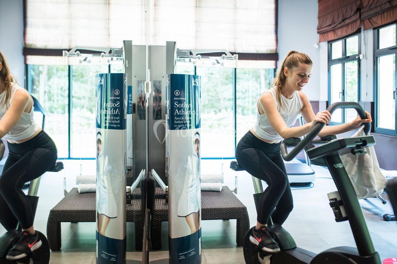 Une femme s'entraîne sur un vélo d'appartement dans une salle de sport lumineuse avec des miroirs.