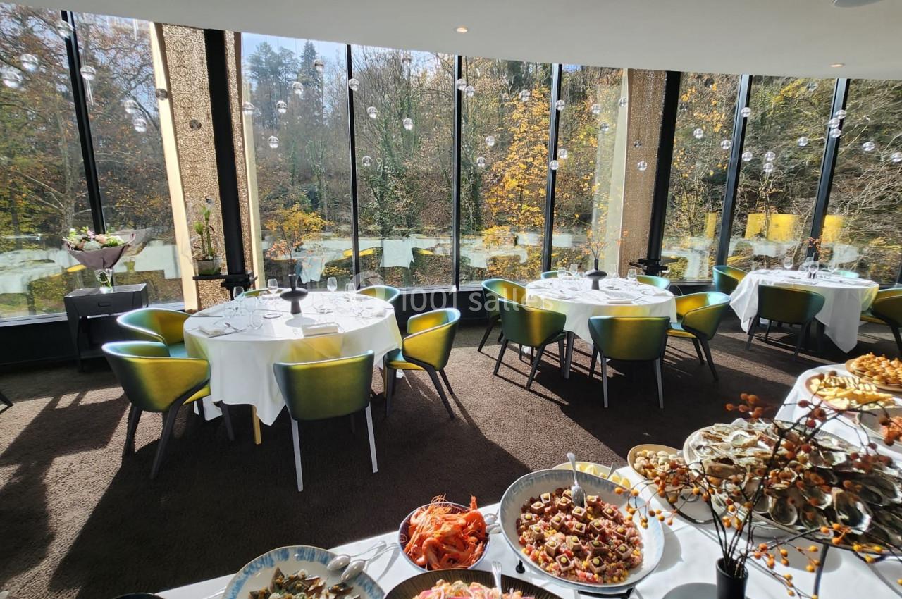 Salle de restaurant lumineuse avec de grandes baies vitrées donnant sur un paysage automnal, tables dressées et buffet.