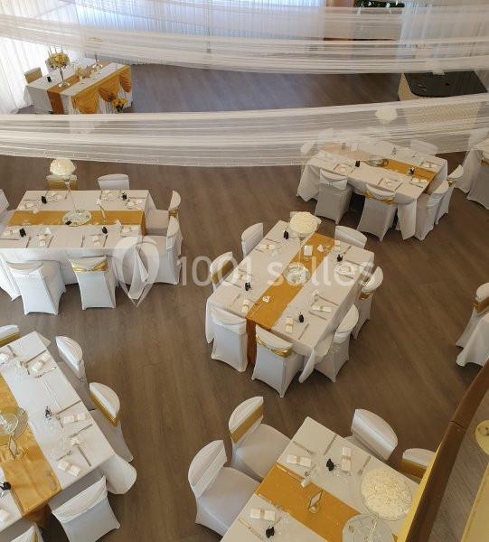 Salle de réception décorée avec des tables rondes et rectangulaires, nappes blanches et chemins de table dorés.