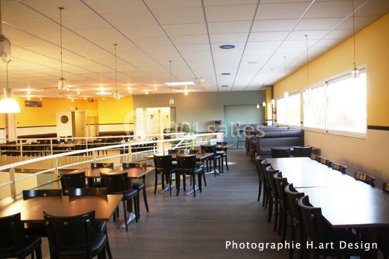 Salle de restaurant lumineuse avec tables et chaises en bois sombre, éclairage suspendu et grandes fenêtres.