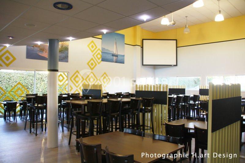 Salle lumineuse avec tables et chaises en bois, décorée de motifs jaunes et de photos murales.
