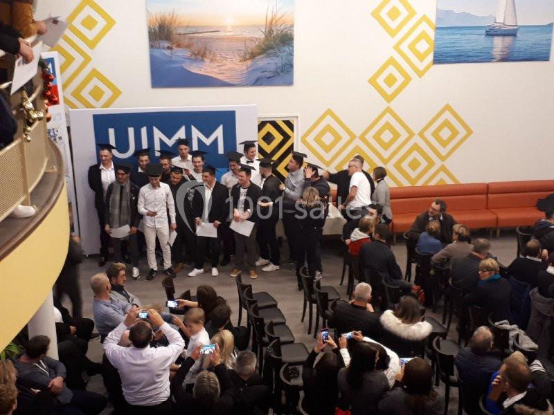 Un groupe de personnes posant devant un panneau ’JIMM’ dans une salle avec des spectateurs assis et des décorations murales.