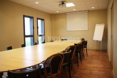 Salle lumineuse avec tables en bois, chaises noires, coin cuisine équipé et grandes fenêtres donnant sur l'extérieur.