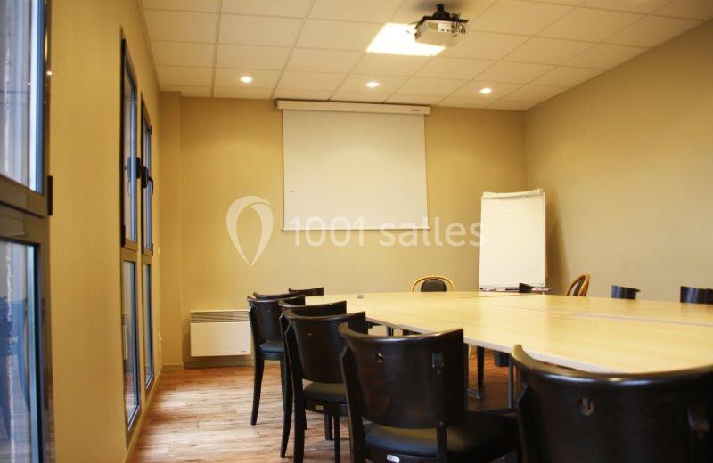 Salle de réunion avec table rectangulaire, chaises noires, écran de projection et paperboard dans un espace lumineux.