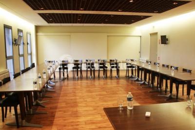 Salle lumineuse avec tables en bois, chaises noires, coin cuisine équipé et grandes fenêtres donnant sur l'extérieur.