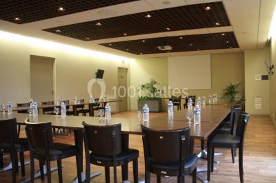 Salle lumineuse avec tables en bois, chaises noires, coin cuisine équipé et grandes fenêtres donnant sur l'extérieur.