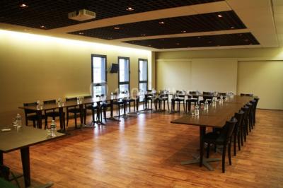 Salle lumineuse avec tables en bois, chaises noires, coin cuisine équipé et grandes fenêtres donnant sur l'extérieur.