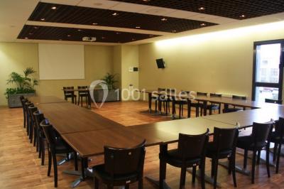 Salle lumineuse avec tables en bois, chaises noires, coin cuisine équipé et grandes fenêtres donnant sur l'extérieur.