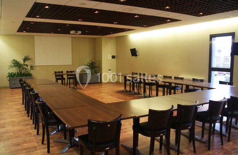 Salle de réunion vide avec tables disposées en U, chaises noires, sol en bois et écran de projection au mur.