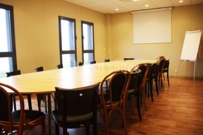 Salle lumineuse avec tables en bois, chaises noires, coin cuisine équipé et grandes fenêtres donnant sur l'extérieur.