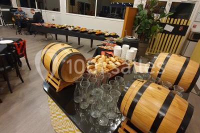 Salle lumineuse avec tables en bois, chaises noires, coin cuisine équipé et grandes fenêtres donnant sur l'extérieur.