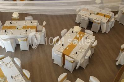 Salle lumineuse avec tables en bois, chaises noires, coin cuisine équipé et grandes fenêtres donnant sur l'extérieur.