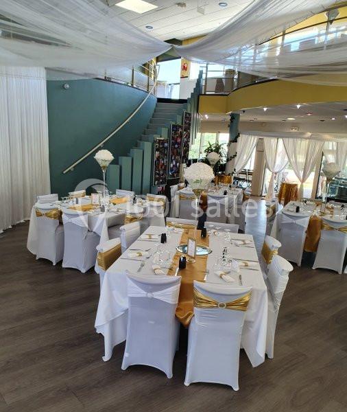 Salle de réception décorée avec des tables rondes, nappes blanches, chemins de table dorés et chaises assorties.